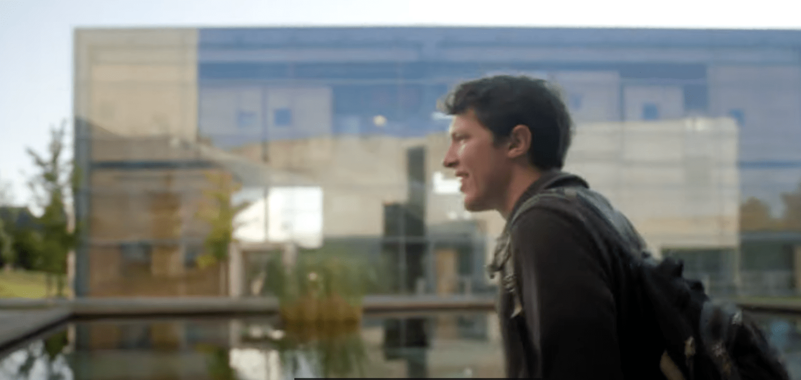 Still from video showing student Patrick Newcombe walking in front of a glass box building.