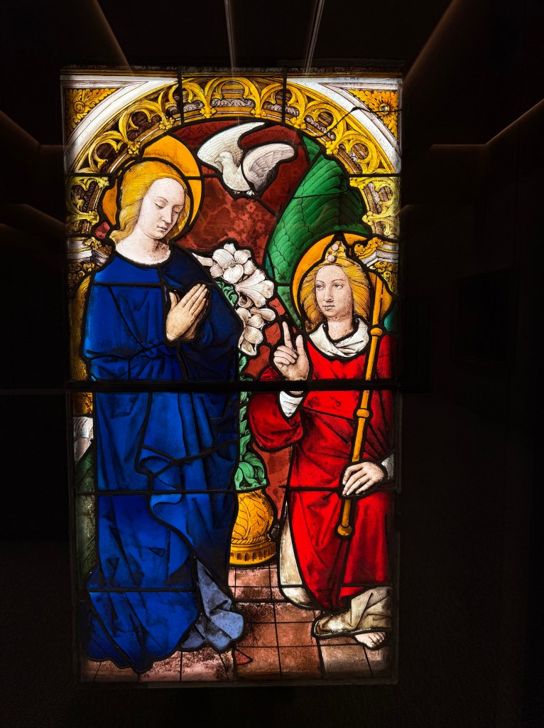 A white dove hovers between Mary, in a blue gown and holding her hands in prayer, and an angel, robed in red with index finger extended.