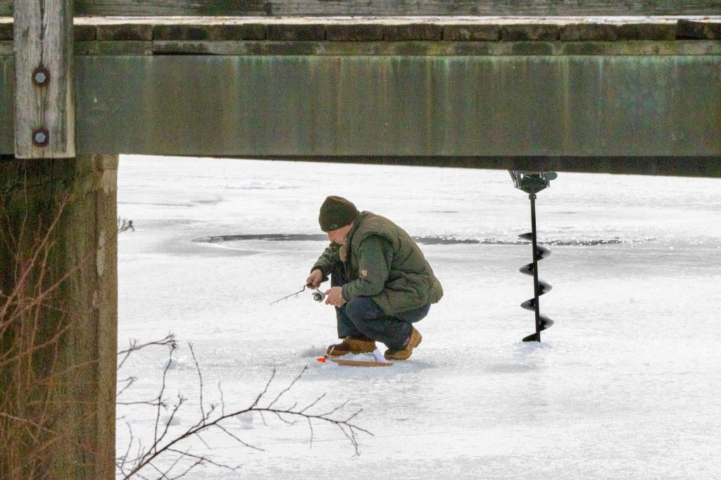 Man ice-fishing on frozen lake adjusts what appears to be a fishing reel as he squats on the ice. An auger is behind him, sticking into the ice.