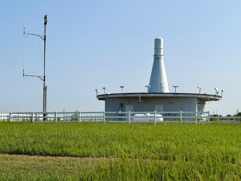 Aviation beacon on hilltop. It in a circular building and looks vaguely like a bowling pin. A tall pole with two horizontal antennae stands nearby.